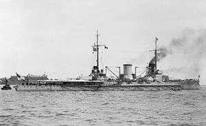 A large warship sits motionless off shore, light gray smoke drifts lazily from its forward smoke stack.