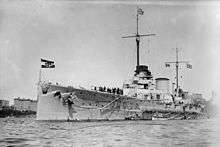 A large warship sits in harbor, with two smaller boats alongside. High-rise buildings are seen in the distance.