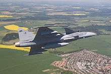 Grey jet aircraft banking right over rural area with residential housing. The background is mostly green with yellow areas.