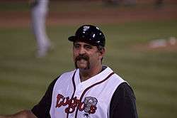 A smiling, mustachioed man in a white-and-black baseball jersey and black baseball helmet