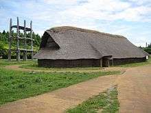 A long very basic wooden house with thatched roof. Behind the house there is a wooden three-storied structure with open floors supported by large pillars.
