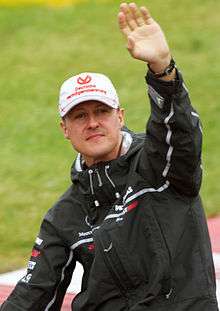 A man in a black jacket and silver cap with his left arm held aloft