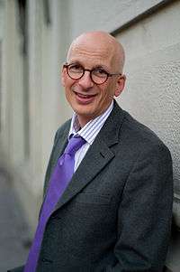 Picture of a white, bald-headed person in his 50s with brown-eyes (looking directly forward) and wearing a black shirt. The person's face is taken behind a red-orange background.