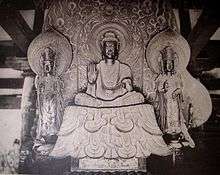Front view of a central figure sitting cross-legged on a raised platform which is flanked by two smaller standing statues. The central figure has the palm of his right hand turned to the front. The attendants look identical, pointing upwards with their right hand and their left hand lifted halfway, touching the thumb with the middle finger. Each of the three statues has a halo.