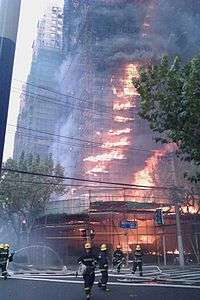 Several firefighters walk near scaffolding that is wrapped around a dark structure with flames shooting out.