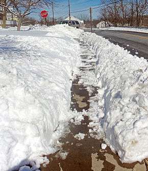 A sidewalk at the bottom of a crudely cut trench through the snow. Some snow on it is melting in the sun.