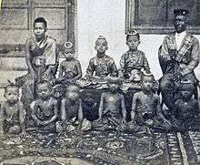 Sepia tinted photo of seated woman and older man separated by three children sitting on a bench, with several children sitting on the floor in front of them