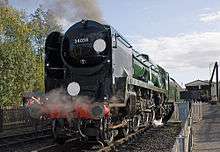 A front view of another 4-6-2 steam locomotive with a tender hauling carriages away from a station. The locomotive is one of the preserved rebuilt examples, and is newly restored.