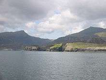 Grey hills back green moors beyond a grey body of water. A small island is separated from the cliffs of the shore by a deep chasm.