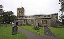 A long stone church seen from the south with a clerestory and a west tower surmounted by a battlemented parapet and a saddleback roof