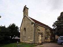 A small simple, stone church seen from the southwest, showing a bellcote, buttresses, and a round-arched doorway and west window, each flanked by columns