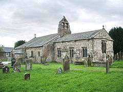 A low stone church with a double bellcote at the junction of the chancel and nave