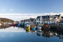 Still blue water in the foreground with numerous small coloured boats along a harbour and buildings against a blue sky in the background