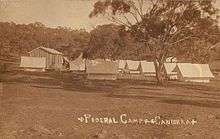 Single wooden cabin and many white tents in open dusty field with single tree in foreground