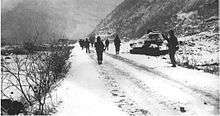 A line of soldiers walking past a destroyed tank