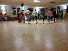 A small circle of dancers with one foot raised hold hands while several people look on.