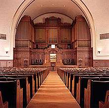 Facing the viewer are the backs of many rows of pews, with a carpeted aisle in the center. At the front of the room is a very large, wooden structure, which fills the far wall.