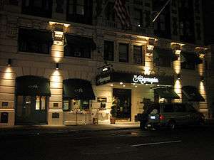 Spotlights accentuate the hotel's stone facade and arched store windows with awnings. A large awning covers the entrance to the hotel.