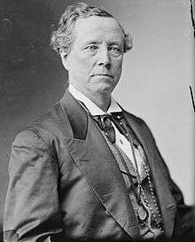 A man with curly, graying hair wearing a white shirt, and a dark jacket, vest, and bowtie