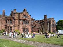 A three-storey house with bays containing mullioned and transomed windows, and multiple gables.  On the lawned area in front of the house are many people presumably enjoying an open day.