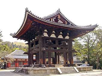 Small wooden house with a hip-and gable roof. The structure is open from all sides and a large bell is hanging in the center.