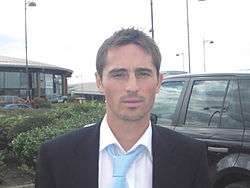 The head and shoulders of a man, wearing a white shirt, blue tie and black blazer