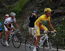 A man in yellow clothes, with a yellow helmet and yellow handgloves, riding a bicycle. Just behind him is another cyclist, in blue clothes.