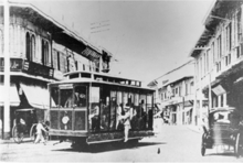 An electric trolley with a man hanging off one side rounds a corner of a street lined by two-story stores and horse-drawn kalesas.