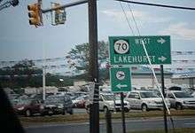 An intersection in a business area with a green sign reading Route 70 west Lakehurst with two right arrows