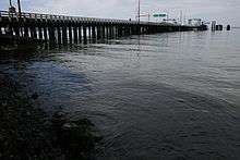 Water is meeting the shore in the foreground of the picture while a pylon based bridge is in the far left corner, which is the ferry dock