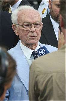 An older gentleman wearing glasses and who is bald except for a crescent of white hair at the back of his head is standing in the middle of a crowd wearing a light blue suit and dark blue tie. A man in a brown shirt whose back is turned to the camera is pointing a blue microphone with the number one at the older man.