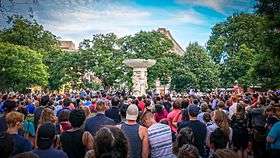 Hundreds of people stand together listening to a man speaking from an outdoor stage. In the center of the crowd is a large statue with a paper sign affixed to it with tape; the sign reads: "D.C. Loves You Orlando".