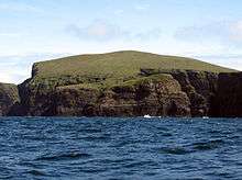 A green hill ends in dark brown cliffs over blue water.