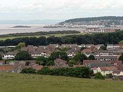 Multiple houses and other buildings around a bay into which a pier projects. On the background are hills.