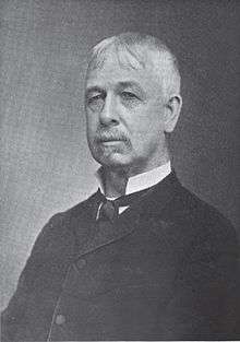 A white-haired man wearing a high-collared white shirt, black tie, and black jacket