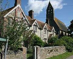 A row of cottages, partially obscured by vegetation. In the background is the bell tower of a much large building.